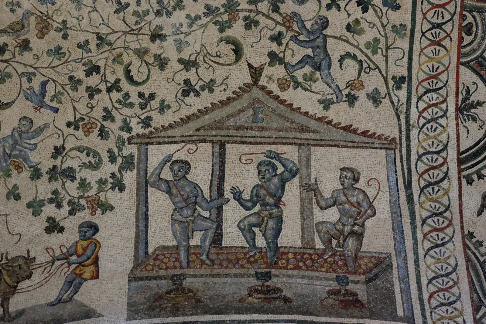 mosaico de la vendimia en una bóveda: bajo un lagar con frontón, tres figuras pisan la uva y el mosto cae a tres tinajas; alrededor, vides con racimos y pájaros