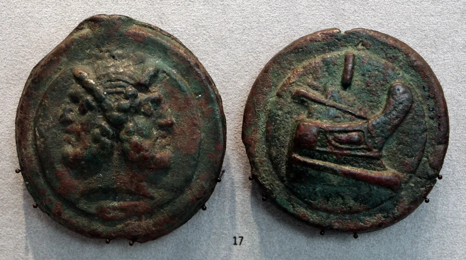 los dos lados del as romano de bronce fundido, en una vitrina: la cabeza doble y barbada de jano y la proa de una nave